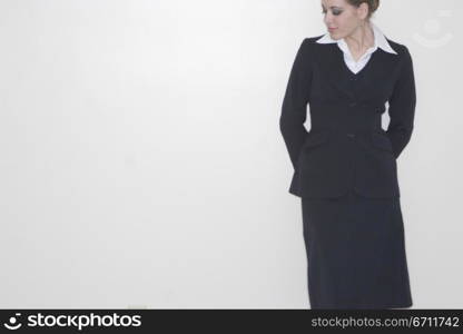 businesswoman standing alone against a white wall with her hands behind her looking down