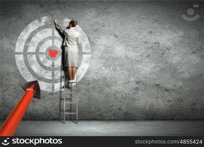 Businesswoman standing on ladder and pointing at target on wall
