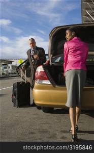 Businesswoman unloading her suitcase from a car trunk with a businessman standing in front of her