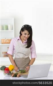 Businesswoman using laptop while chopping vegetable in kitchen