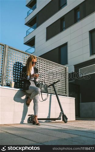 Businesswoman using mobile sitting on a wall with her scooter next. Businesswoman using mobile with her scooter next
