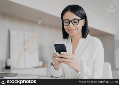 Businesswoman using phone mobile corporate app in office, reading message, chatting, getting good news. Smiling female in glasses ordering, shopping online in workplace, looking at smartphone.. Businesswoman in glasses using phone mobile apps in office, reads good news message, chatting online