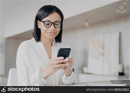 Businesswoman using phone mobile corporate app in office, reading message, chatting, getting good news. Smiling female in glasses ordering, shopping online in workplace, looking at smartphone.. Businesswoman in glasses using phone mobile apps in office, reads good news message, chatting online
