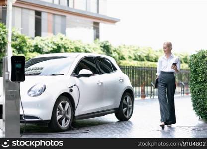 Businesswoman using tablet, walking while recharging her electric vehicle with charging station at public car parking. Progressive lifestyle of technology and ecological concern by EV car. Progressive businesswoman using tablet at EV charging station.