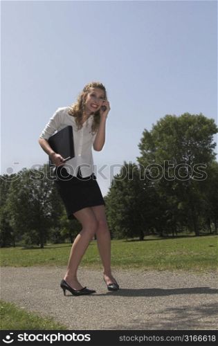 Businesswoman walking in the park with her laptop under her arm and calling on her mobile.