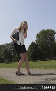 Businesswoman walking in the park with her laptop under her arm and calling on her mobile.