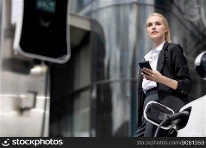 Businesswoman wearing black suit using smartphone, leaning on electric car recharge battery at charging station in city residential building with condos and apartment. Progressive lifestyle concept.. Progressive businesswoman with mockup smartphone with EV at charging station.