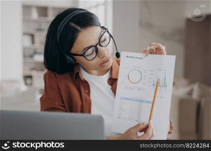 Businesswoman wearing glasses and headset presenting business project during online video conference on laptop, showing document with statistics. Female coach explain charts while elearning.. Businesswoman in headset presenting business project on video conference on laptop, showing document