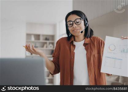Businesswoman wearing glasses, headset, talking by corporate video call showing infographic. Professional female business coach holding document with graphs, discussing project online.. Businesswoman in glasses, headset, discuss business project online by video call showing infographic