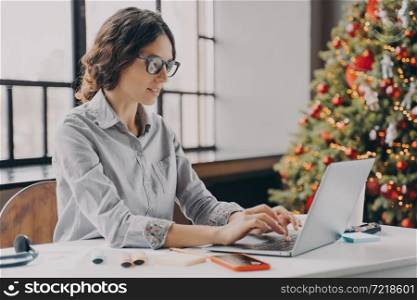 Businesswoman wearing glasses sitting in office near xmas tree and working on laptop during the holiday season, typing on computer keyboard. Hispanic female employee spending Christmas at work. Businesswoman wearing glasses sitting in office near xmas tree and working on laptop
