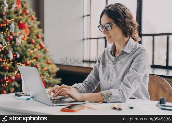 Businesswoman wearing glasses sitting in office near xmas tree and working on laptop during the holiday season, typing on computer keyboard. Hispanic female employee spending Christmas at work. Businesswoman wearing glasses sitting in office near xmas tree and working on laptop