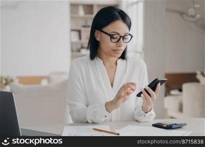 Businesswoman wearing glasses using mobile phone apps for business time management sitting at laptop. Focused serious female manager answers messages in social networks on smartphone at desk.. Businesswoman in glasses using mobile phone apps for business time management sitting at laptop