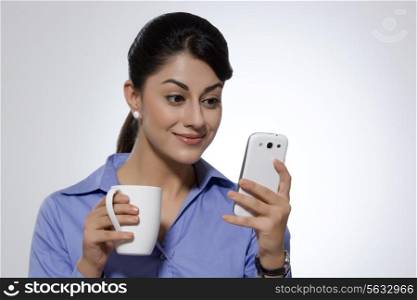 Businesswoman with coffee mug smiling while reading text message on smart phone over gray background
