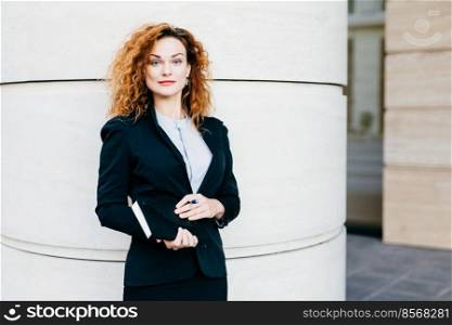 Businesswoman with curly light hair, red painted lips, wearing white blouse, black jacket and skirt, holding notebook with pen, going to write necessary notes. People, success and business concept