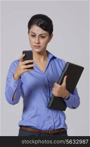 Businesswoman with laptop reading text message on smart phone against gray background