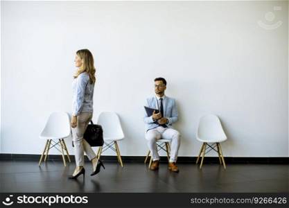 Businesswoman with suitcase passing by young man sitting at chair in the waiting room with a folder in hand before an interview