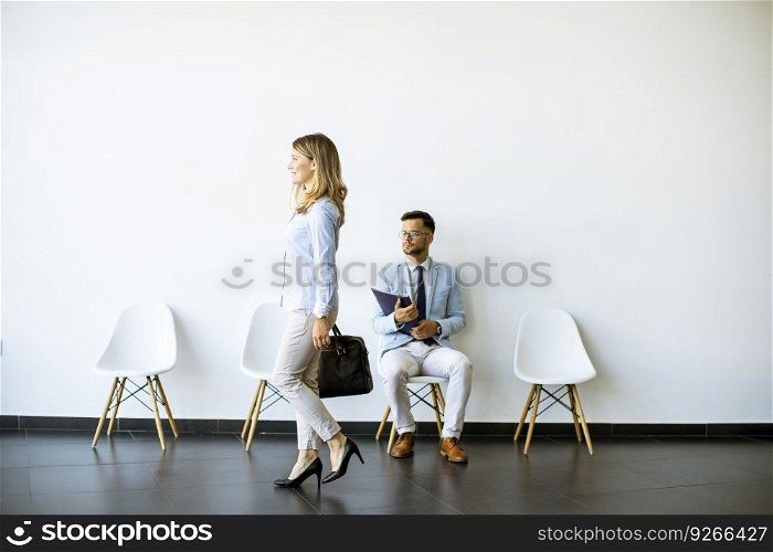 Businesswoman with suitcase passing by young man sitting at chair in the waiting room with a folder in hand before an interview