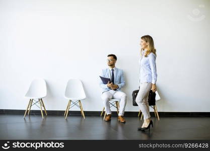 Businesswoman with suitcase passing by young man sitting at chair in the waiting room with a folder in hand before an interview