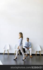 Businesswoman with suitcase passing by young man sitting at chair in the waiting room with a folder in hand before an interview