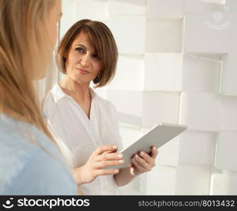 Businesswoman with tablet looking at her colleague while standing against of modern white wall.. Portrait of brunette woman with tablet looking at her colleague