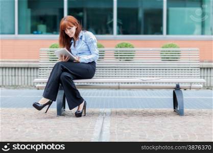 Businesswoman with tablet pc is sitting outside on a metal bank and looking towards the camera
