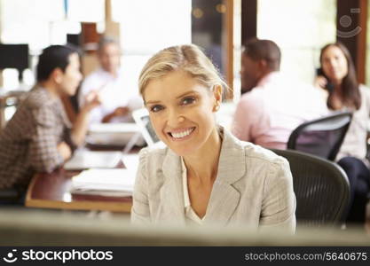 Businesswoman Working At Desk With Meeting In Background