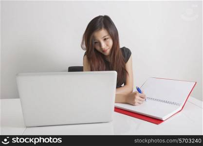 Businesswoman writing in diary while looking at laptop in office