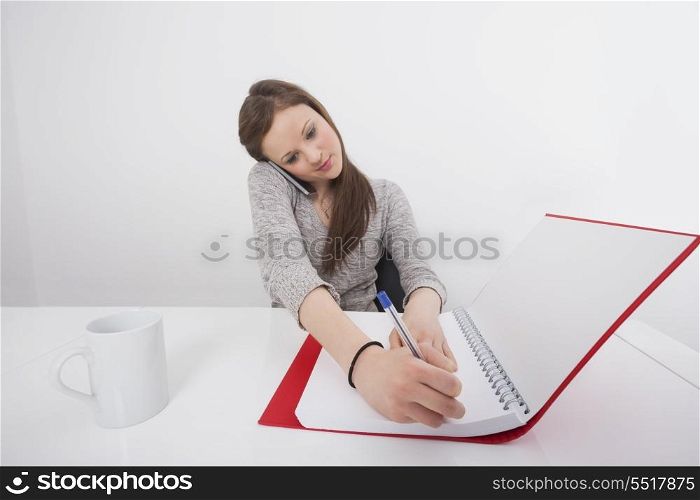 Businesswoman writing notes while answering smart phone in office