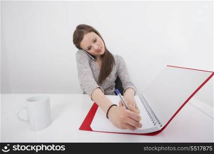 Businesswoman writing notes while answering smart phone in office