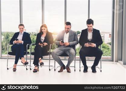 Businesswomen and businessmen using mobile phone while waiting on chairs in office for job interview. Corporate business and human resources concept.. Businesswomen and businessmen using mobile phone.