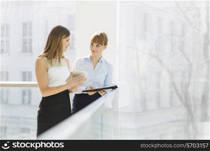 Businesswomen discussing over tablet PC while standing by railing in office