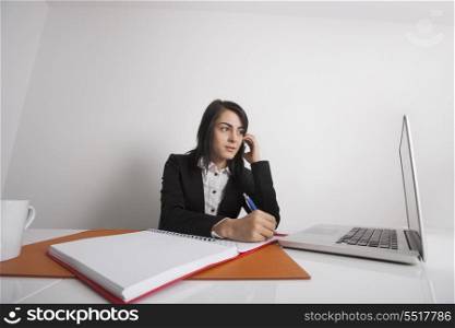 Businesswomen using cell phone while writing notes from laptop at office desk