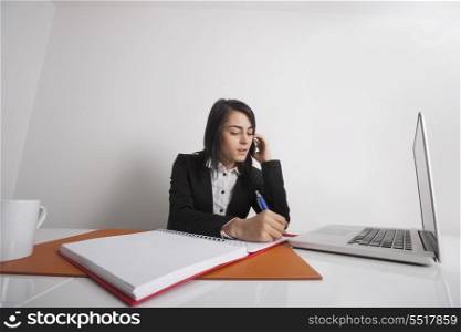 Businesswomen writing notes while using cell phone at office desk