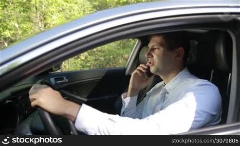 Busy at work. Positive businessman sitting in luxury car holding steering wheel with one hand and talking on smartphone during business trip in summer. Confident young entrepreneur communicating on cell phone with colleague while driving car.