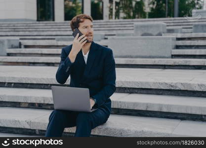 Busy bearded male entrepreneur busy working with statistics data discusses performance via mobile phone call looks away uses laptop computer dressed in black formal suit sits outdoor on stairs. Busy male entrepreneur busy working with statistics data discusses performance via mobile phone