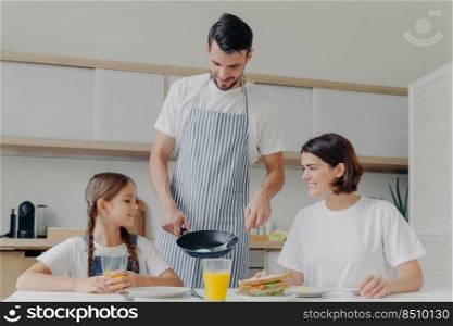 Busy caring father prepares delicious breakfast for wife and daughter, wears apron, puts fried eggs on plate. Cheerful mother and child talk to each other while sit at kitchen table, have meal