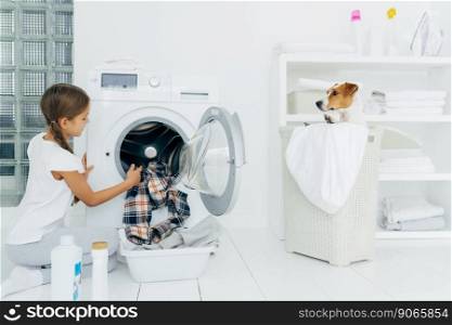 Busy child does laundry work, empties washing machine, cleaned clothes in basin uses detergents, little pedigree dog in basket. Modern household device at home. Female kid helps with family chores
