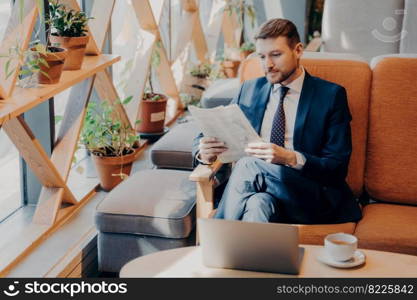 Busy corporate employee dressed in blue suit sitting on comfortable chair in cozy cafe after ordering coffee, taking break from work, reading newspaper with open laptop in front of him. Busy corporate employee reading newspaper in cafe