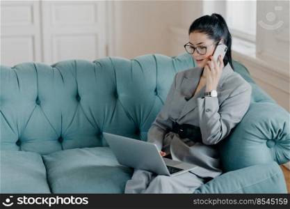 Busy female designer has cell phone conversation, sits in front of opened laptop computer on comfortable sofa, discusses new project. Businesswoman dressed in formal suit works remotely from home