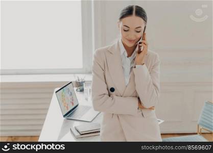 Busy female director in formal suit having cell phone conversation, talking on phone and discussing new project with copartner. Businesswoman working at light office with laptop in wooden desk. Successful business owner talking on mobile phone