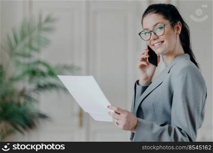 Busy female entrepreneur has telephone conversation, busy with accounting work, looks through paper documents, checks information, wears spectacles and formal costume, poses indoor, cozy interior
