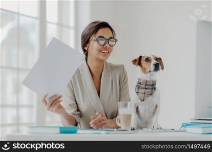 Busy female entrepreneur works with papers, prepares business report, concentrated in monitor of computer, dressed formally, dog sits near, pose at desktop with notepads around. Pet helps working