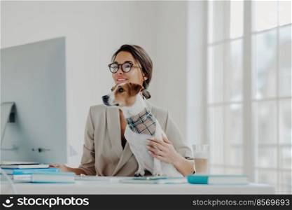 Busy female teacher wears formal clothes and spectacles conducts online lessons for students works at computer poses in coworking space with dog, busy working online. Businesswoman at desktop