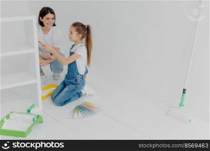 Busy girl and her smiling mother paint drawer in white color, use paintbrush or roller, surrounded with color s&les, pose in white empty room, think about house decoration. Domestic work concept