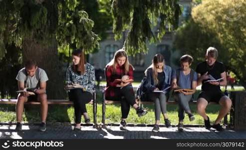 Busy group of college students reading books and lecture notes while sitting on the bench in the park on campus. Frontal view. Serious teenage friends studying together on fresh air while preparing for exams.