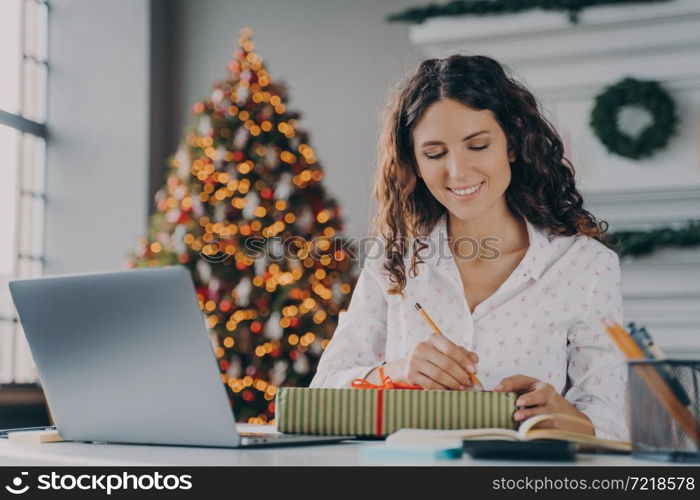 Busy holidays. Young beautiful happy Italian business woman signs prepared Xmas gift box for clients or colleagues with joyful smile while sitting at desk in home office decorated with New Year tree. Young beautiful happy Italian business woman signs prepared Xmas gift box for client in office