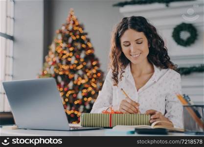 Busy holidays. Young beautiful happy Italian business woman signs prepared Xmas gift box for clients or colleagues with joyful smile while sitting at desk in home office decorated with New Year tree. Young beautiful happy Italian business woman signs prepared Xmas gift box for client in office