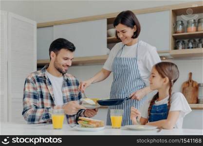 Busy housewife holds frying pan, gives prepared meal to husband and daughter, have delicious breakfast together, sit at kitchen table with glass of juice and sandwich. Happy family eat at home
