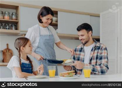 Busy housewife holds frying pan, gives prepared meal to husband and daughter, have delicious breakfast together, sit at kitchen table with glass of juice and sandwich. Happy family eat at home