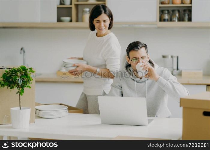 Busy husband works on laptop computer and drinks coffee, wears spectacles and casual sweatshirt, wife busy unpacking belongings, carries dishes, pose in kitchen during moving day. Family and mortgage